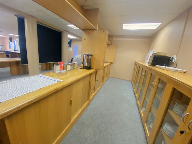 A functional kitchenette area featuring built-in wooden cabinetry, glass-fronted display cupboards, and a laminate countertop, suitable for office or commercial breakroom use.
