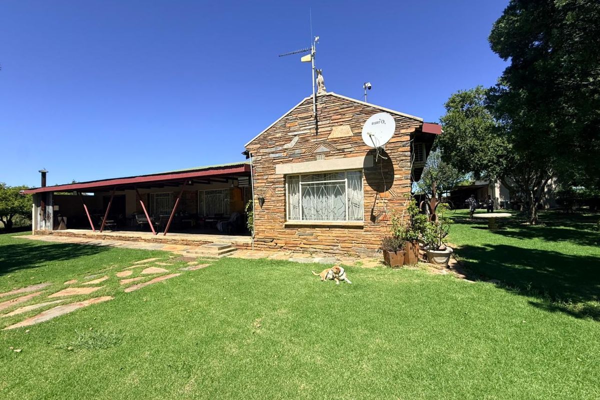 Rear view of a stone-clad farmhouse featuring a spacious covered patio, expansive lawn, and established garden surroundings.