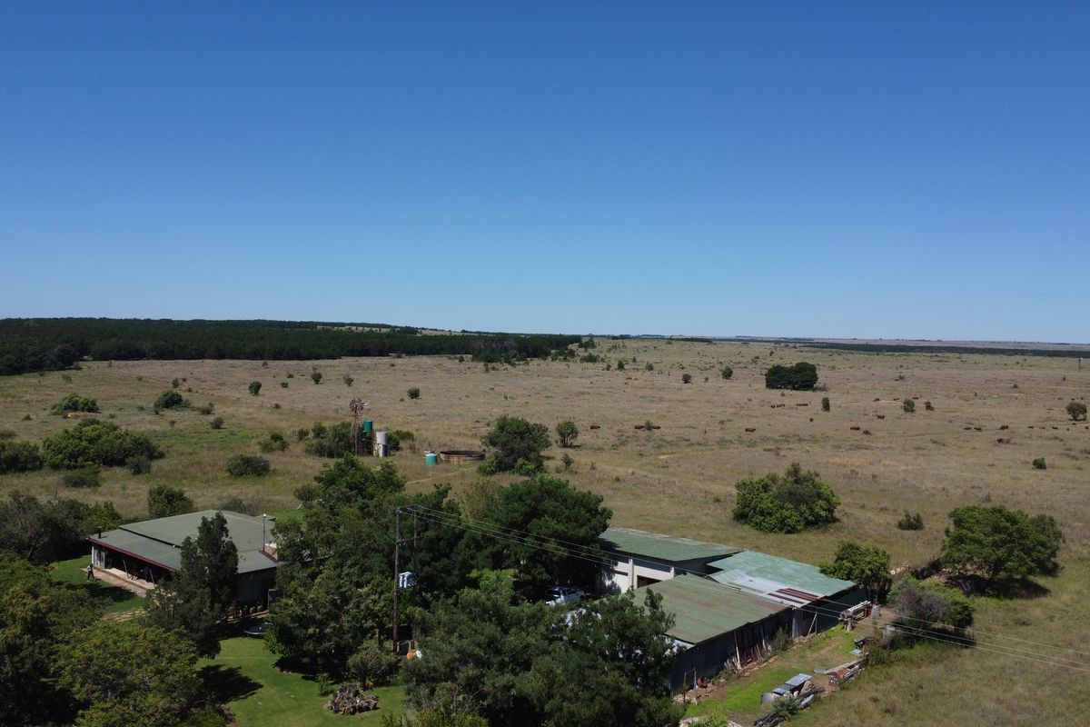 Aerial view of a farm property featuring multiple outbuildings, a windmill, and expansive surrounding grazing land under a clear blue sky.