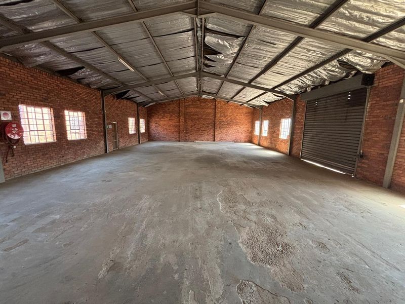 Spacious industrial warehouse unit featuring exposed brick walls, concrete flooring, steel roof trusses, and a large roller shutter door for easy access.