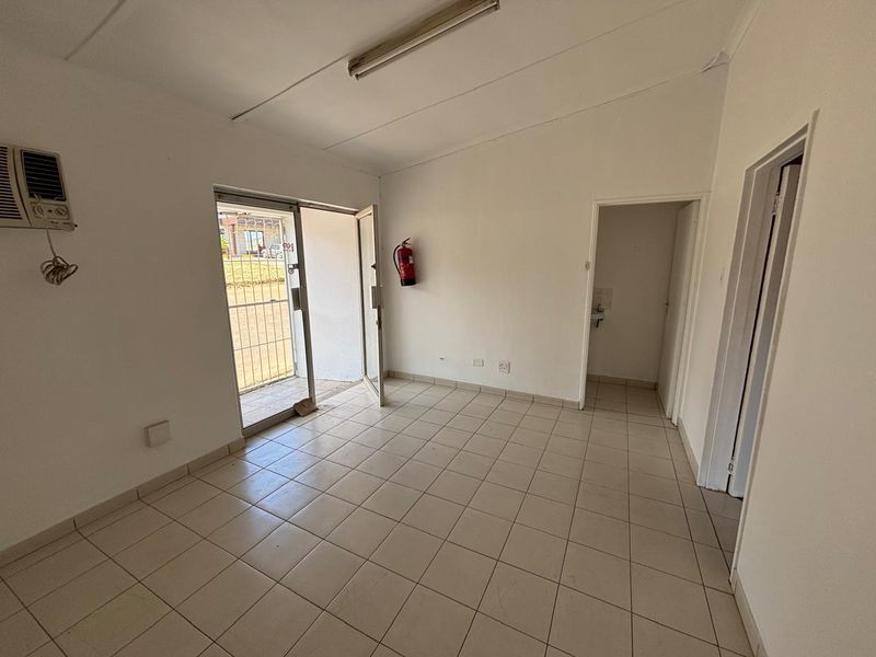 An empty industrial office space featuring tiled flooring, a wall-mounted air conditioning unit, and a security gate leading to the exterior.
