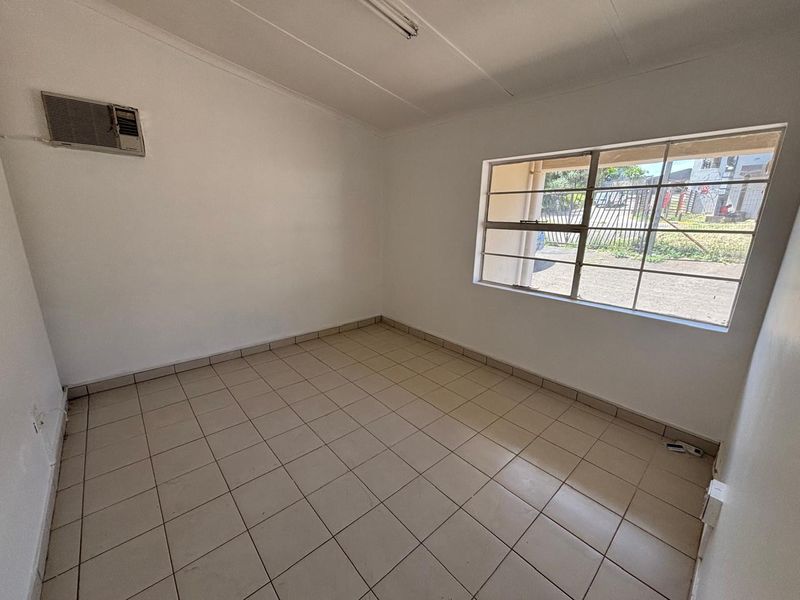 A clean, empty office space featuring tiled flooring, a wall-mounted air conditioning unit, and a window with security bars.