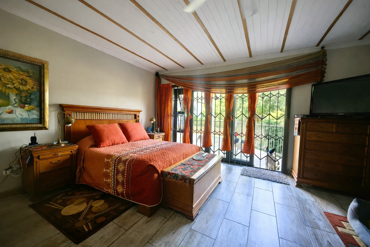 A spacious bedroom featuring wood-look tile flooring, an exposed beam ceiling, and a sliding glass door with security gate leading to the outdoors.
