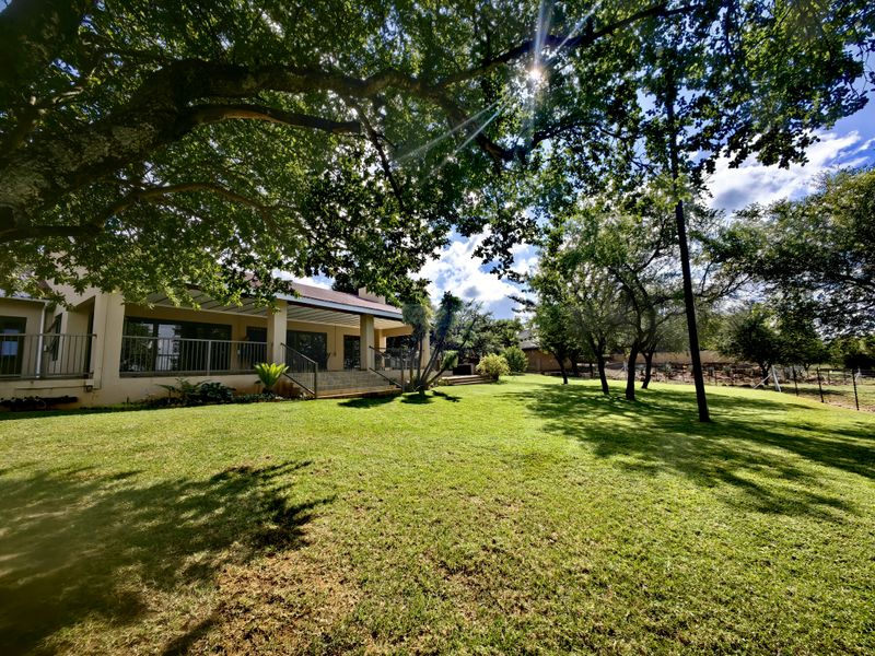 A spacious rear garden featuring a well-maintained lawn, mature trees, and a covered patio area with metal railings and concrete steps.