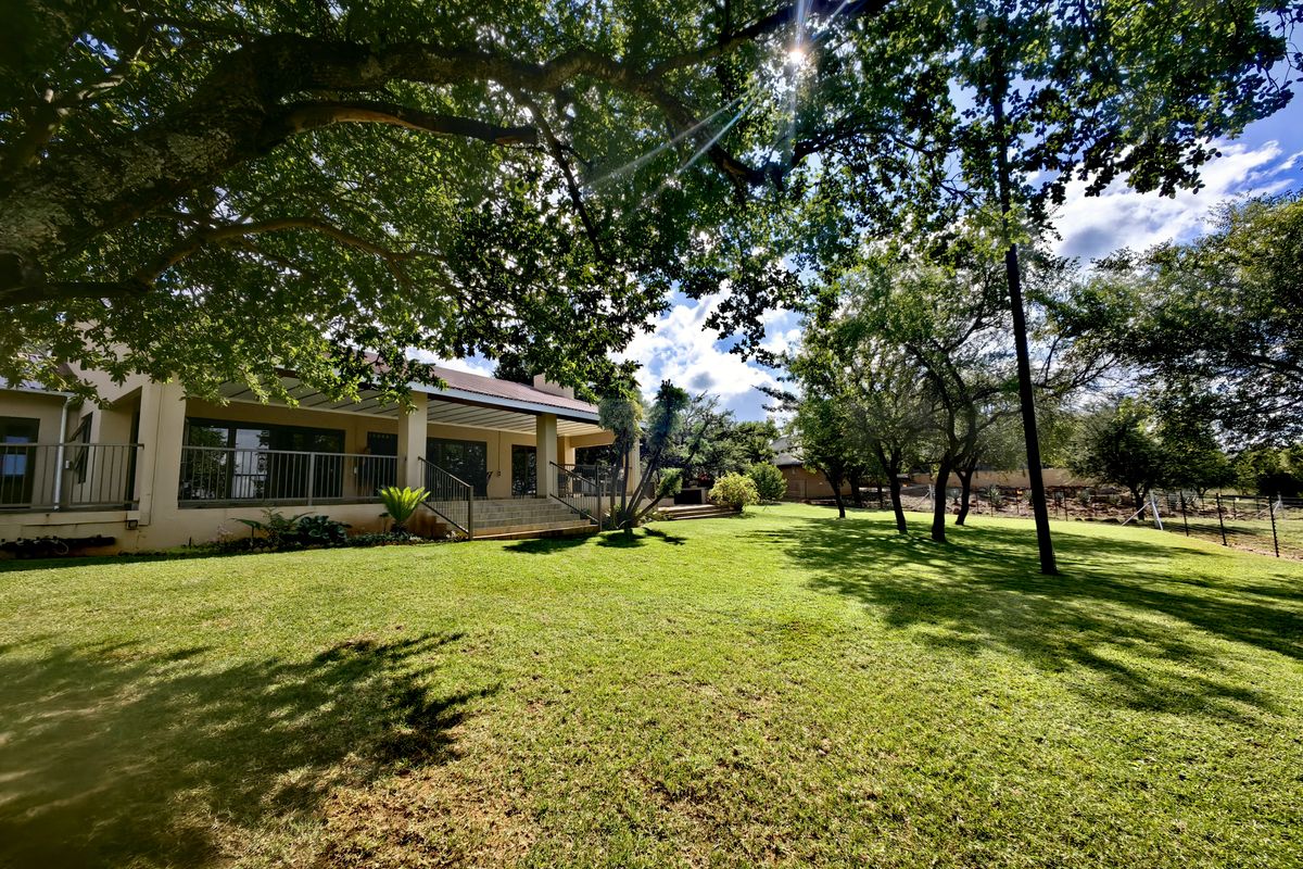 A spacious rear garden featuring a well-maintained lawn, mature trees, and a covered patio area with metal railings and concrete steps.