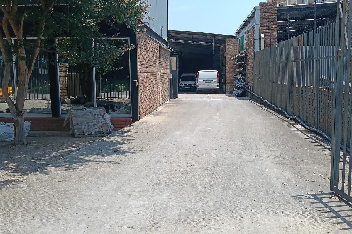 Industrial property exterior featuring a concrete driveway leading to a warehouse entrance, enclosed by perimeter fencing and brick building structures.