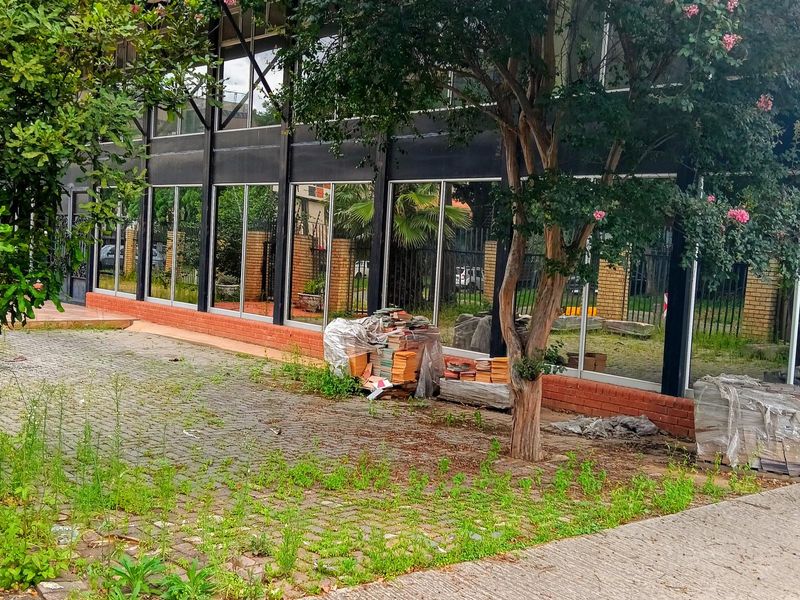 Industrial building exterior featuring large glass panels, black steel framing, and a brick base, set within a paved area with mature landscaping.