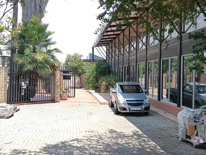 Exterior view of an industrial property featuring a paved driveway, large glass facade with steel framing, and mature landscaping.