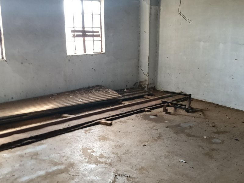 An industrial utility space featuring exposed steel roof trusses, concrete flooring, and steel-framed windows, requiring significant renovation and finishing.