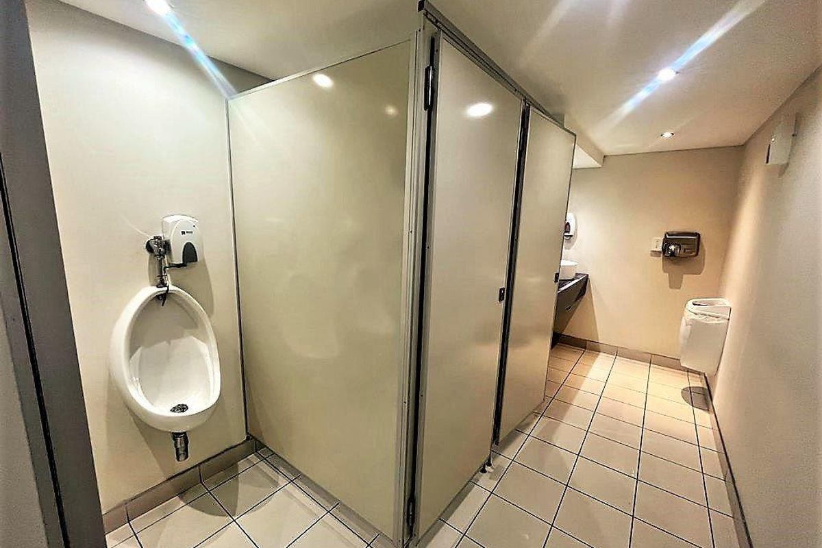 A functional office restroom featuring tiled flooring, multiple toilet cubicles, and wall-mounted urinals with integrated soap dispensers.
