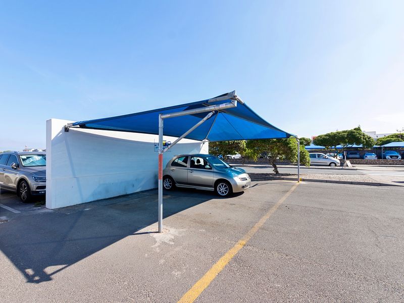 Outdoor parking area featuring a blue shade sail structure and a concrete boundary wall on an asphalt surface.