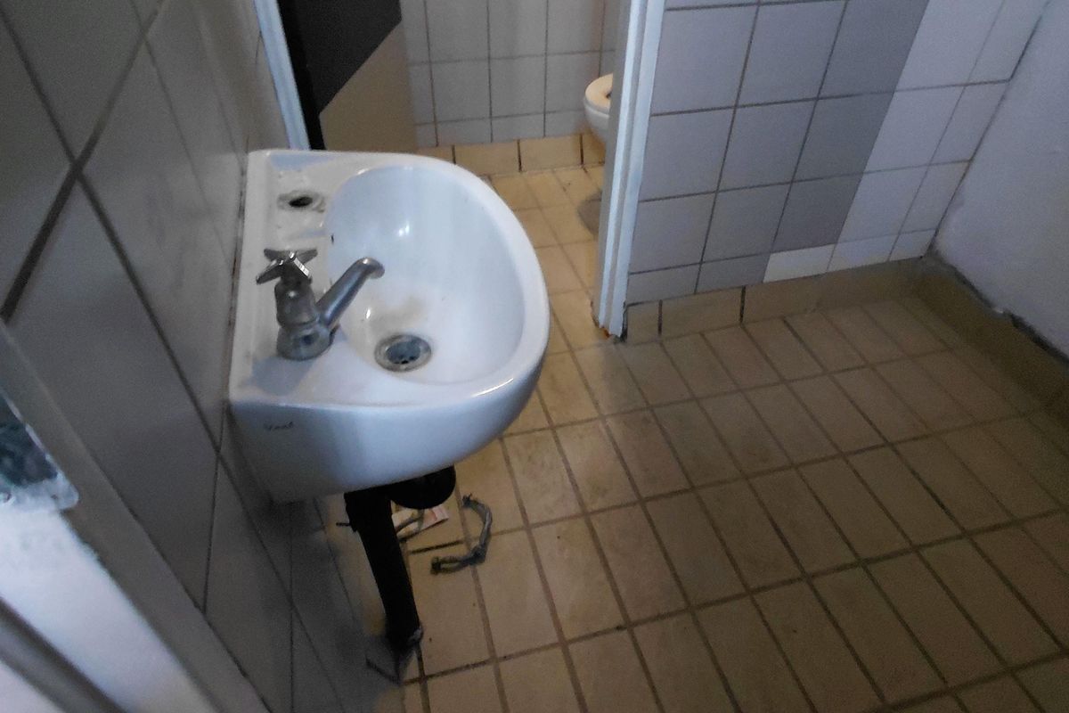 A functional restroom facility featuring a wall-mounted basin, tiled flooring, and wall tiling, requiring maintenance and cosmetic updates.