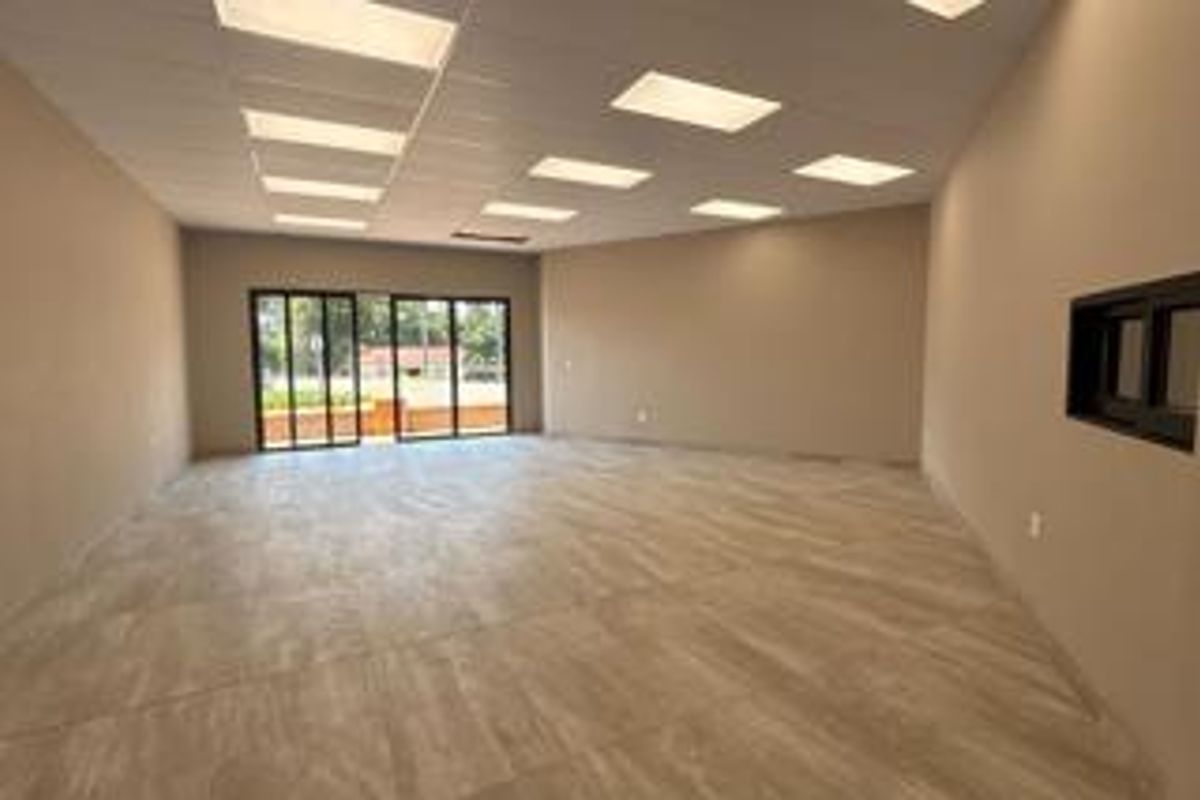 A spacious, vacant office area featuring modern tiled flooring, recessed ceiling lighting, and large sliding glass doors providing natural light and outdoor access.