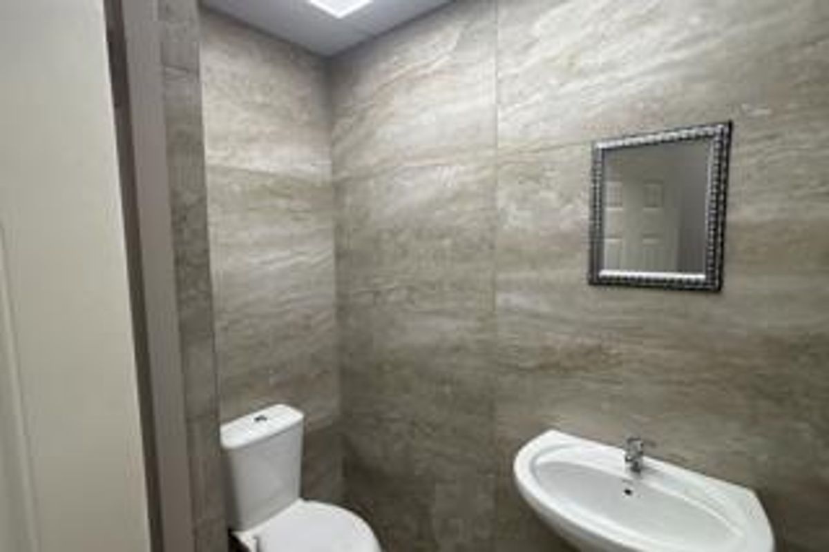 A compact, modern bathroom featuring full-height neutral ceramic wall tiles, a pedestal washbasin, and a standard toilet.