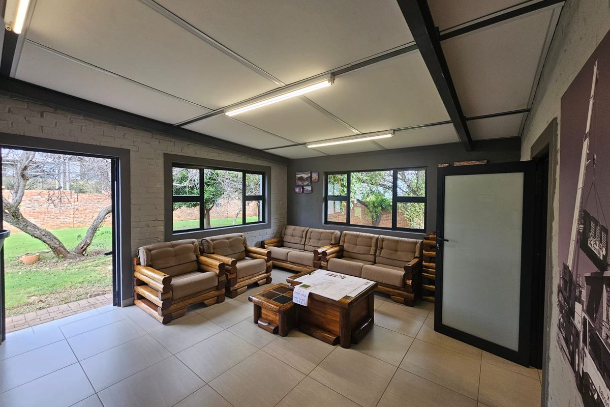 A bright living area featuring tiled floors, exposed steel ceiling beams, and large aluminium windows providing views of the garden.