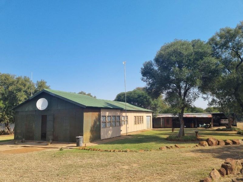 Rear view of a farm property featuring a metal-clad structure with a corrugated roof, set within a spacious garden area with mature trees.