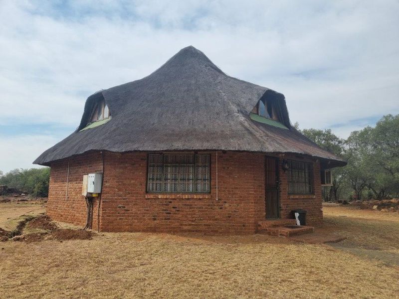A traditional circular face brick farmhouse featuring a thatched roof, dormer windows, and established rural surroundings.