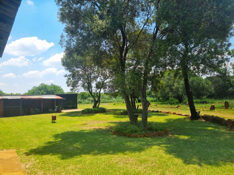 A spacious, well-maintained farm garden featuring mature trees, manicured lawn, stone landscaping elements, and a functional outbuilding in the background.