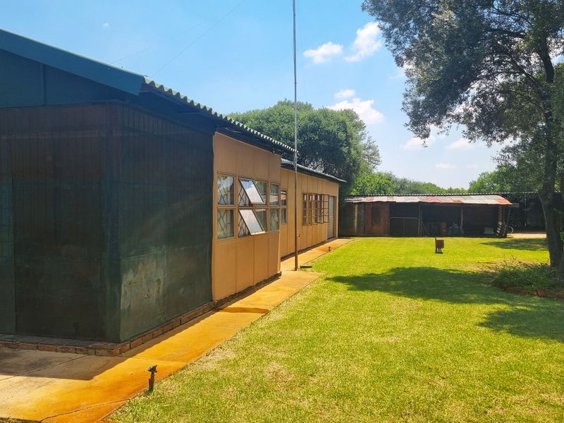 Rear view of the farm property showing the main dwelling with a corrugated roof, concrete walkway, and a separate outbuilding set in a garden.