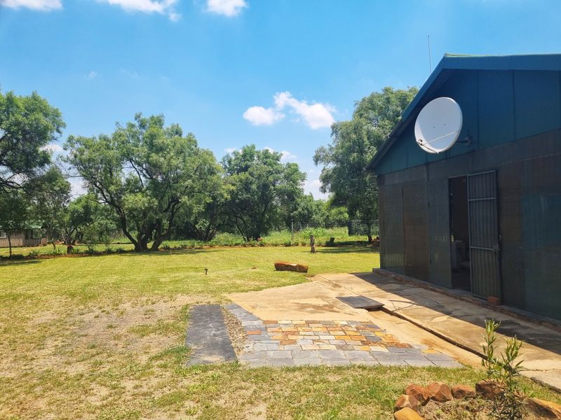 Rear view of a farm building featuring metal cladding, a satellite dish, and a small paved patio area set within a spacious garden.