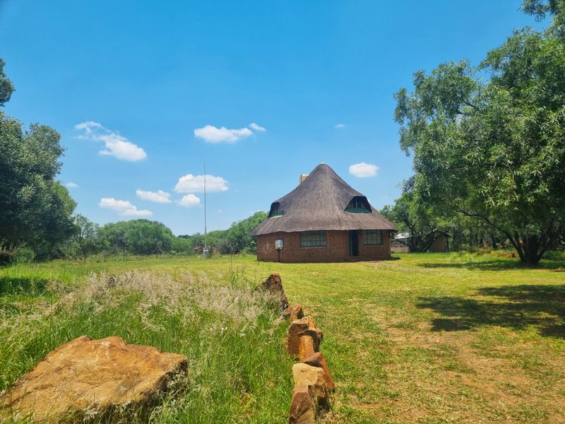 A charming round farmhouse featuring a traditional thatched roof and brick exterior, set within a spacious, natural garden landscape.
