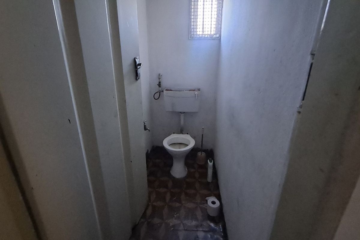 A small, dated bathroom featuring a standard toilet, cistern, and a high-level window with a security grille.