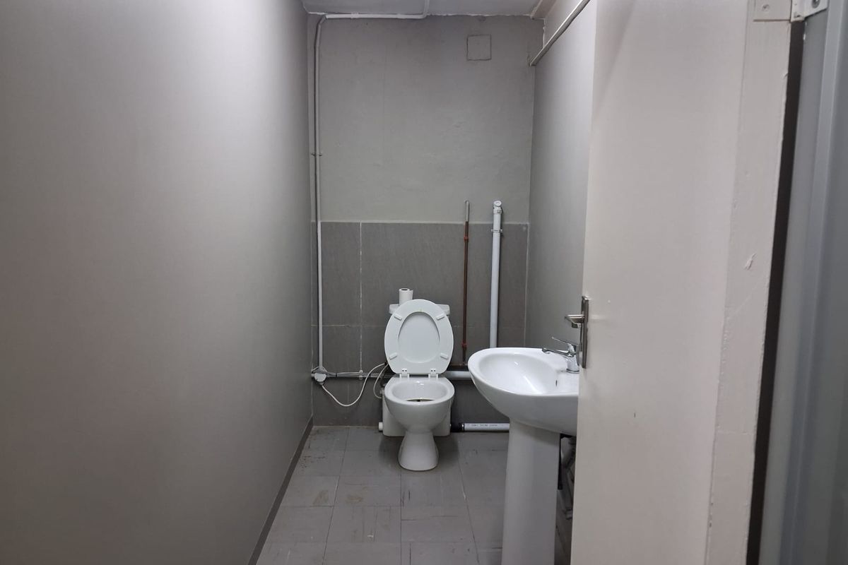 A functional office restroom featuring a standard pedestal basin and toilet with exposed surface-mounted plumbing and tiled flooring.