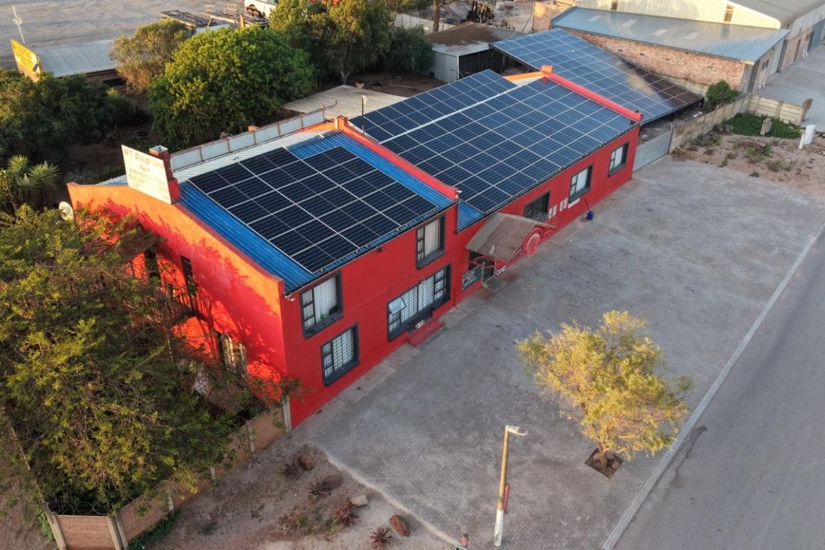 Aerial view of a commercial property featuring a large red building with extensive rooftop solar panel installations and adjacent paved parking area.