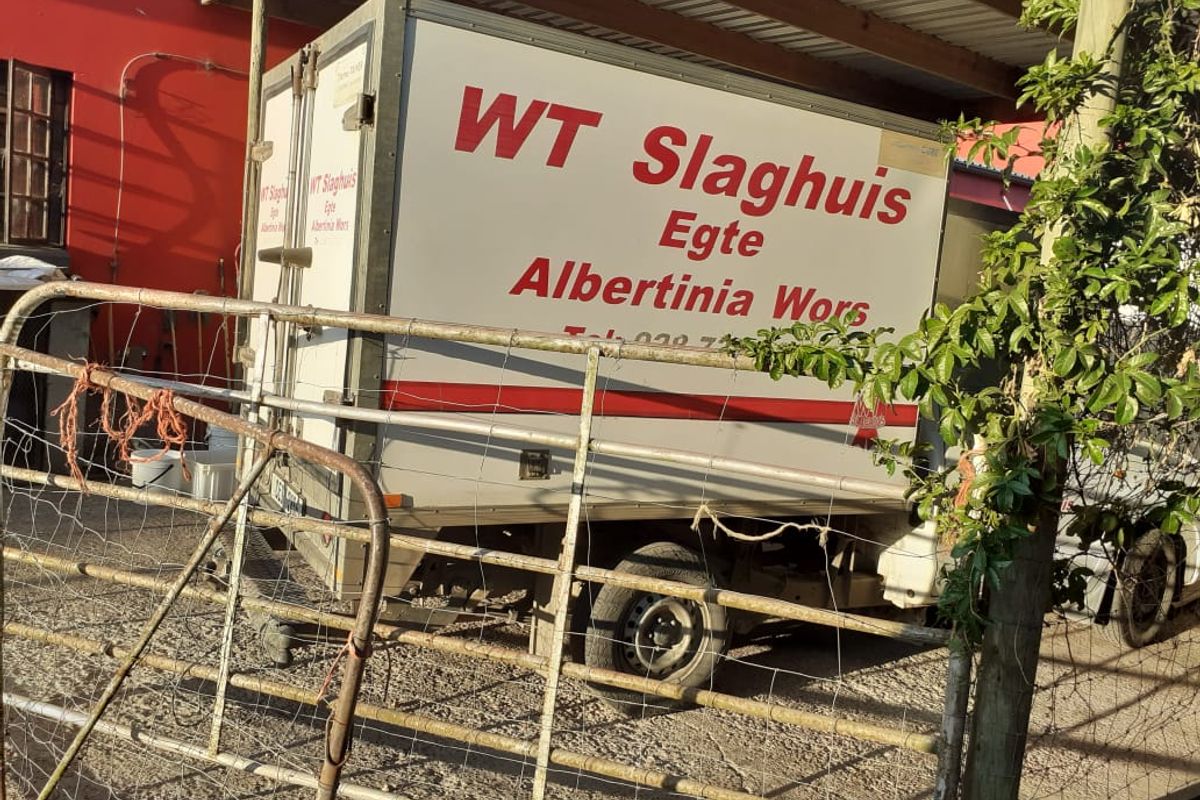 Exterior view of a business property featuring a delivery vehicle parked under a covered structure with metal fencing in the foreground.