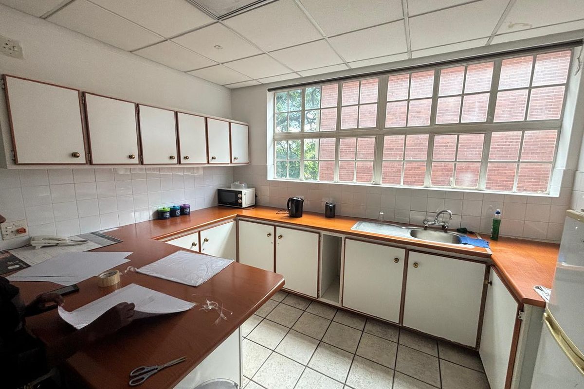 A functional office kitchenette featuring laminate countertops, built-in white cabinetry, a stainless steel sink, and large windows providing natural light.