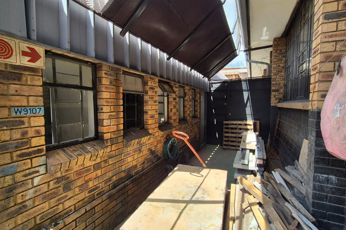 A narrow exterior side passage featuring exposed brick walls, a metal awning, and fire safety equipment, suitable for industrial utility or storage use.