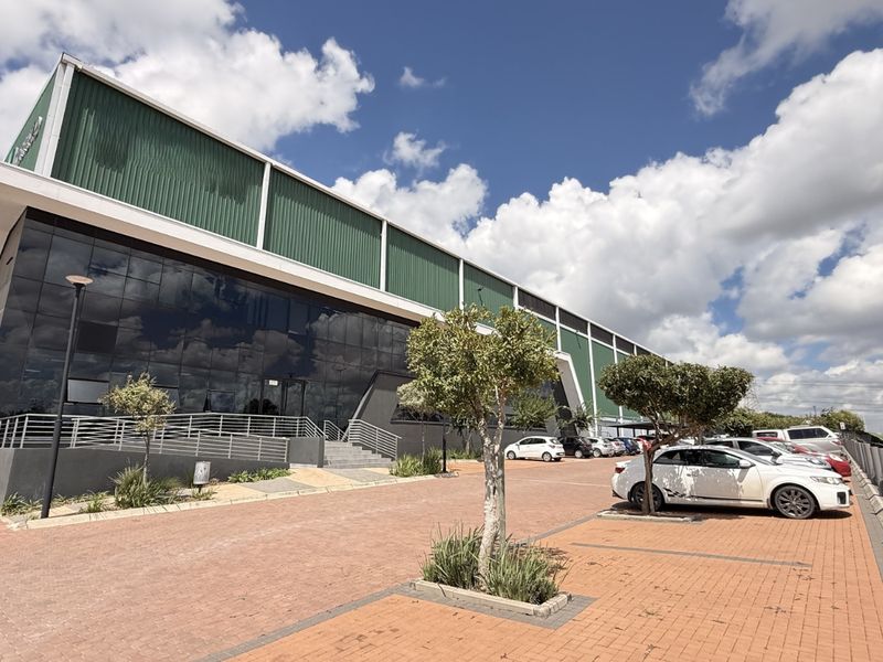 Modern industrial facility featuring a glass facade, green corrugated metal cladding, and an expansive paved parking area with landscaped trees.