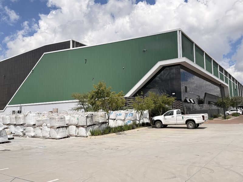 A large industrial warehouse facility featuring green corrugated metal cladding, a modern glass curtain wall facade, and an expansive concrete yard area.