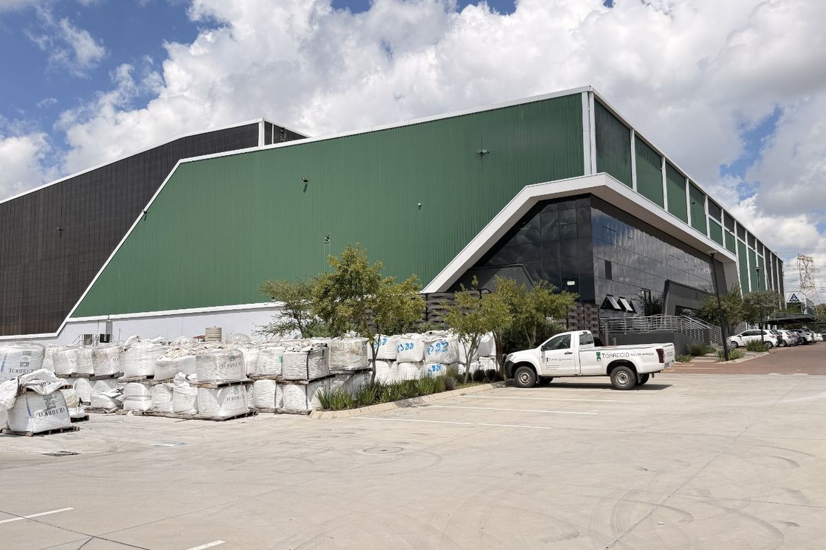 A large industrial warehouse facility featuring green corrugated metal cladding, a modern glass curtain wall facade, and an expansive concrete yard area.