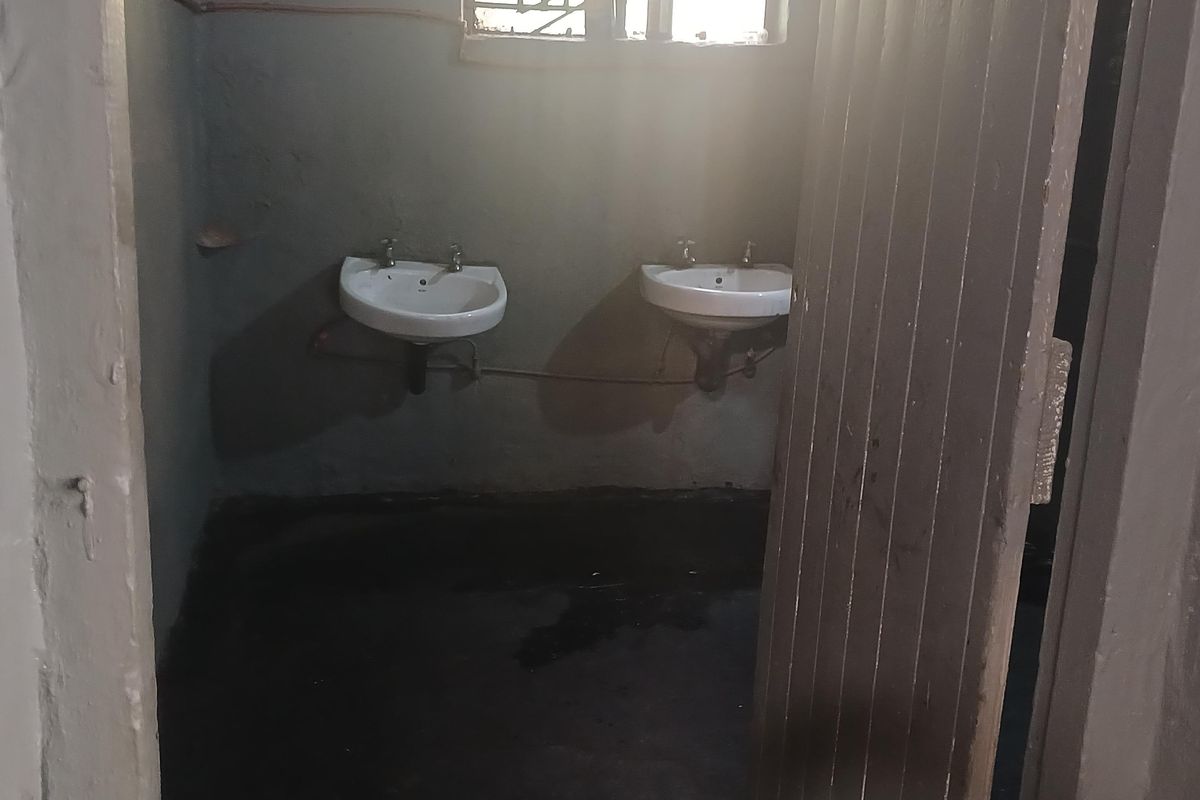 A basic industrial washroom facility featuring two wall-mounted ceramic basins, a small window, and a simple wooden door entrance.