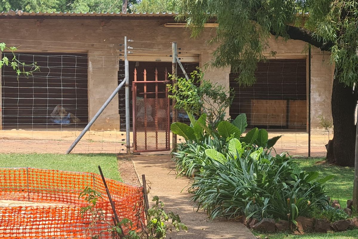 Rear view of a farm outbuilding featuring a concrete path, metal security gate, and electric fencing, set within a garden area.