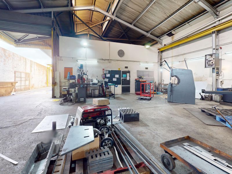 Spacious industrial workshop featuring high ceilings with exposed steel trusses, concrete flooring, and a large roller shutter door for vehicle or equipment access.