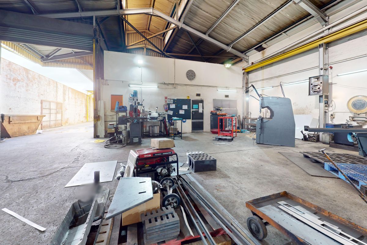 Spacious industrial workshop featuring high ceilings with exposed steel trusses, concrete flooring, and a large roller shutter door for vehicle or equipment access.
