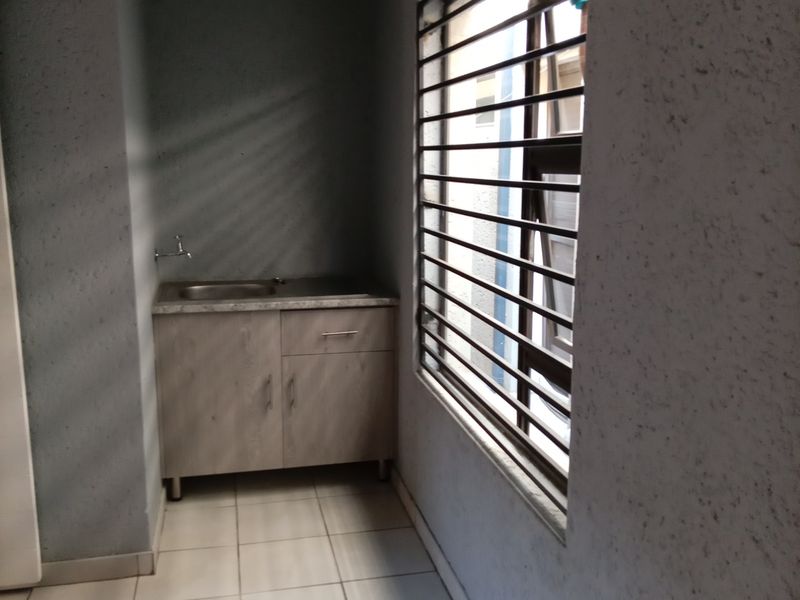 A compact scullery or utility area featuring built-in cabinetry, a stainless steel sink, tiled flooring, and a window with security bars.
