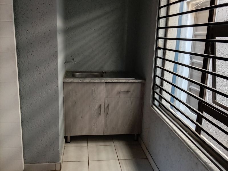 A compact scullery or utility area featuring built-in cabinetry, a stainless steel sink, tiled flooring, and a window with security bars.