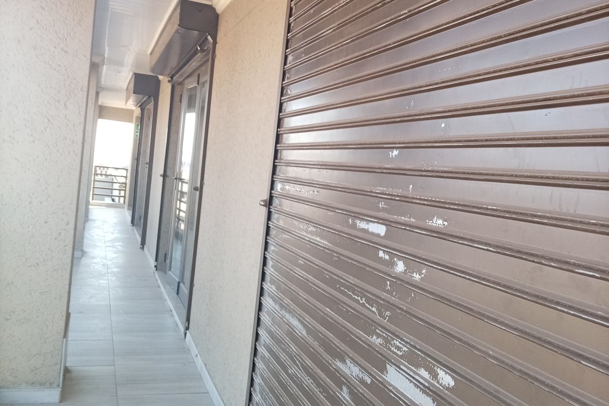 A commercial exterior walkway featuring tiled flooring, recessed ceiling lights, and a metal roller shutter door with visible paint wear.