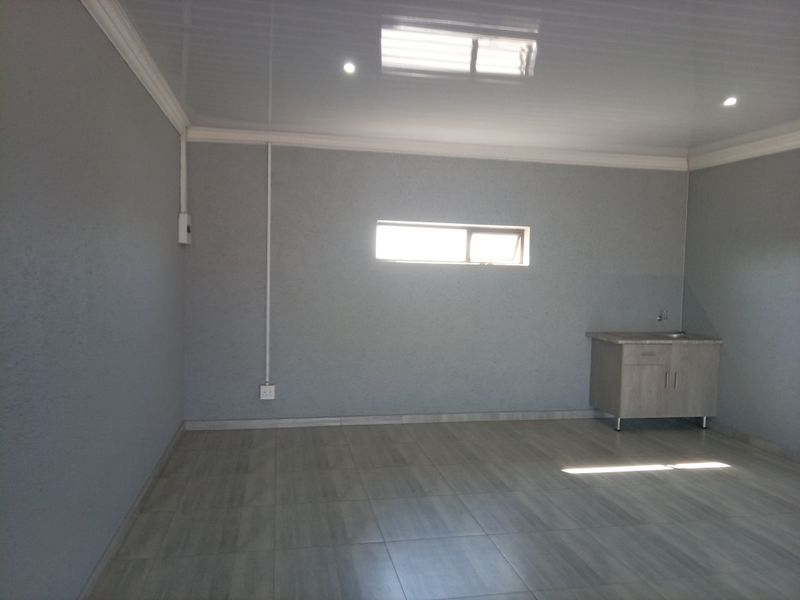 An empty, open-plan commercial space featuring a glossy ceiling, recessed lighting, tiled floors, and a small built-in kitchenette unit with a sink.