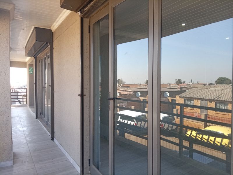 A covered balcony featuring tiled flooring, aluminium sliding doors with integrated security shutters, and a metal balustrade overlooking the surrounding neighbourhood.