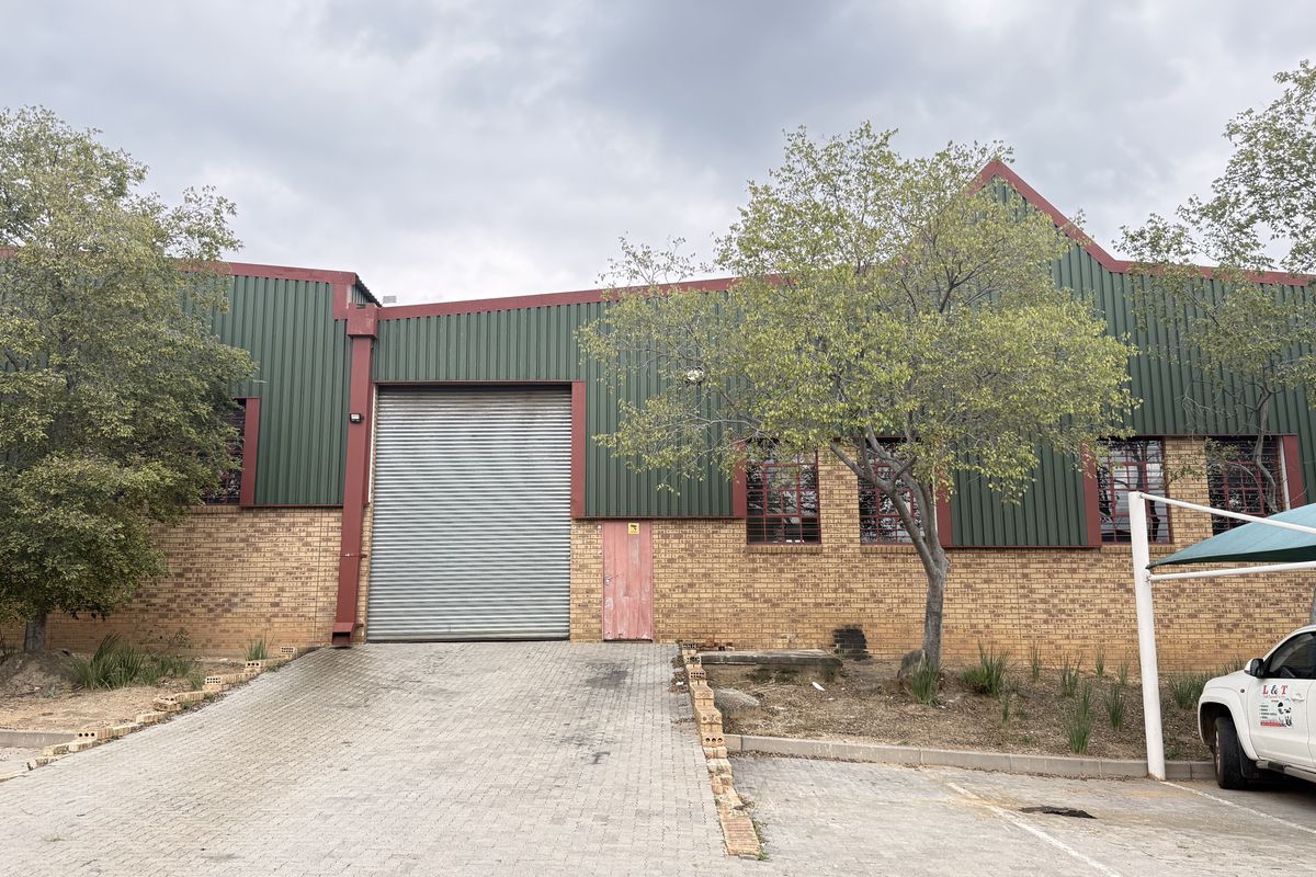 Industrial warehouse exterior featuring a large roller shutter door, brick and metal cladding facade, and a paved driveway with an adjacent shade port.