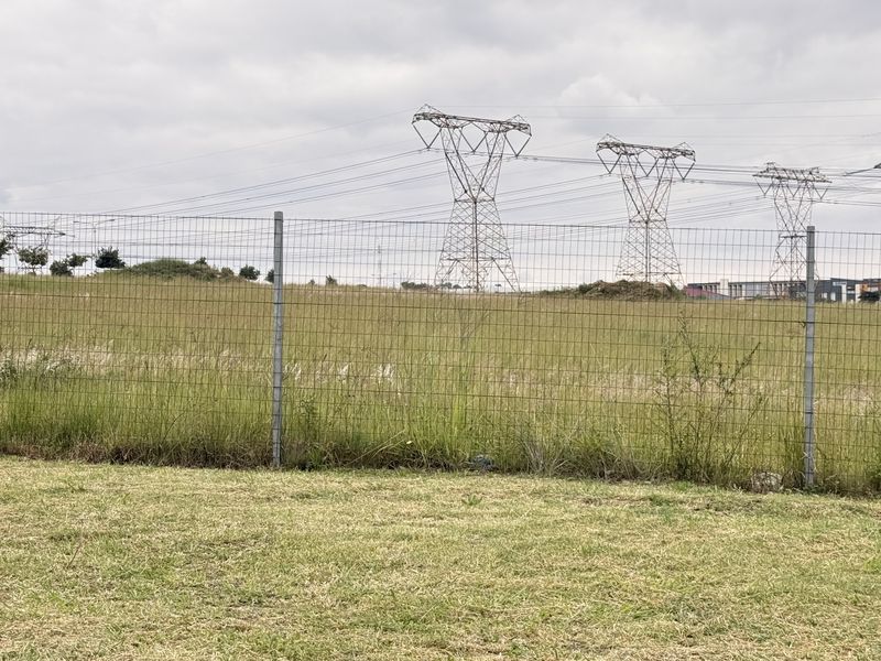 A view of a vacant commercial land plot enclosed by wire mesh fencing, featuring prominent high-voltage electricity pylons in the background.