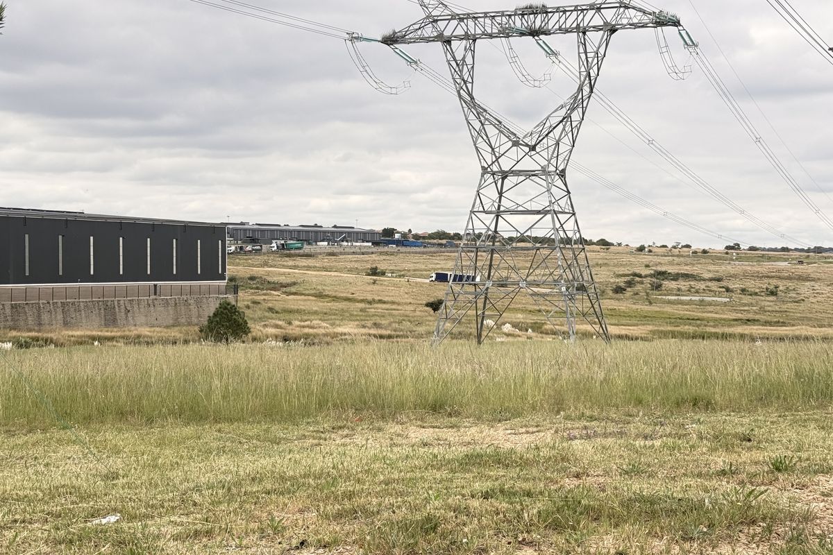 View of vacant commercial land featuring a high voltage transmission tower and adjacent warehouse structure under an overcast sky.