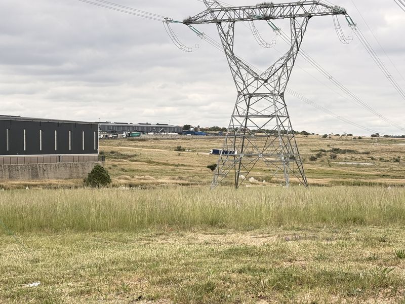 View of vacant commercial land featuring a high voltage transmission tower and adjacent warehouse structure under an overcast sky.