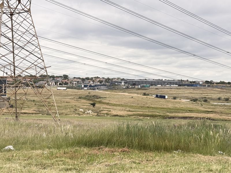 A wide view of vacant commercial land featuring a prominent high-voltage electricity pylon and overhead power lines with distant industrial buildings.