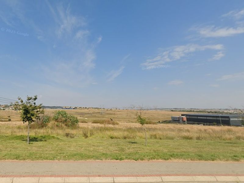 A view of a vacant commercial land plot featuring a paved sidewalk and grass verge, with surrounding industrial development visible in the distance.