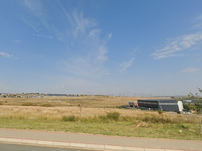 A view of an undeveloped commercial land plot from the adjacent street, featuring a paved sidewalk and surrounding industrial development.